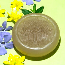 Load image into Gallery viewer, Closeup of a round, pale-colored, glycerin face soap with an embossed tree design on the front and embedded brown specks of rhassoul clay that can be seen,  laying on a bright yellow background with bright yellow and purple flowers, in bright sunlight. 
