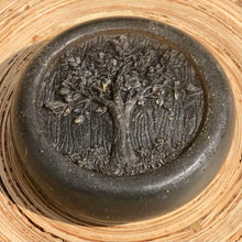 Load image into Gallery viewer, Closeup of a round, grey, glycerin face soap with an embossed tree design on the front and embedded black specks of charcoal and small, brown pieces of hemp that can be seen, laying against blonde wood in the sunlight. 
