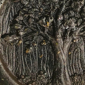 An extreme close-up view of grey face soap with an embossed tree design, and embedded black specks of charcoal and small, brown pieces of hemp that can be seen.