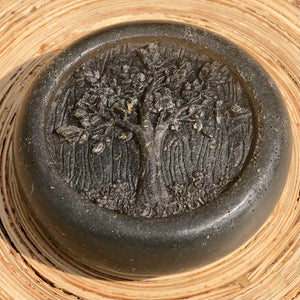 Closeup of a round, grey, glycerin face soap with an embossed tree design on the front and embedded black specks of charcoal and small, brown pieces of hemp that can be seen, laying against blonde wood in the sunlight. 