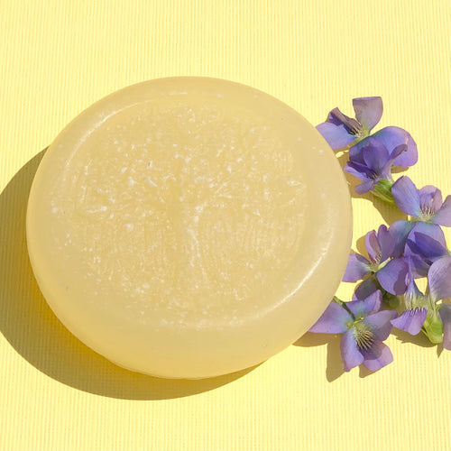 Closeup of a pale yellow, round, glycerin face soap with an embedded tree design, sitting on a bright yellow background, surrounded by small purple flowers on the right side.