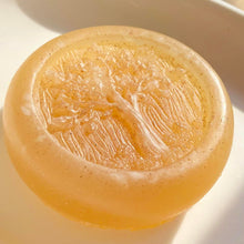 Load image into Gallery viewer, Closeup of a round, pale-colored, glycerin face soap with an embossed tree design on the front and embedded brown specks of rhassoul clay that can be seen, bathed in bright, dappled sunlight against a cream-colored background. 
